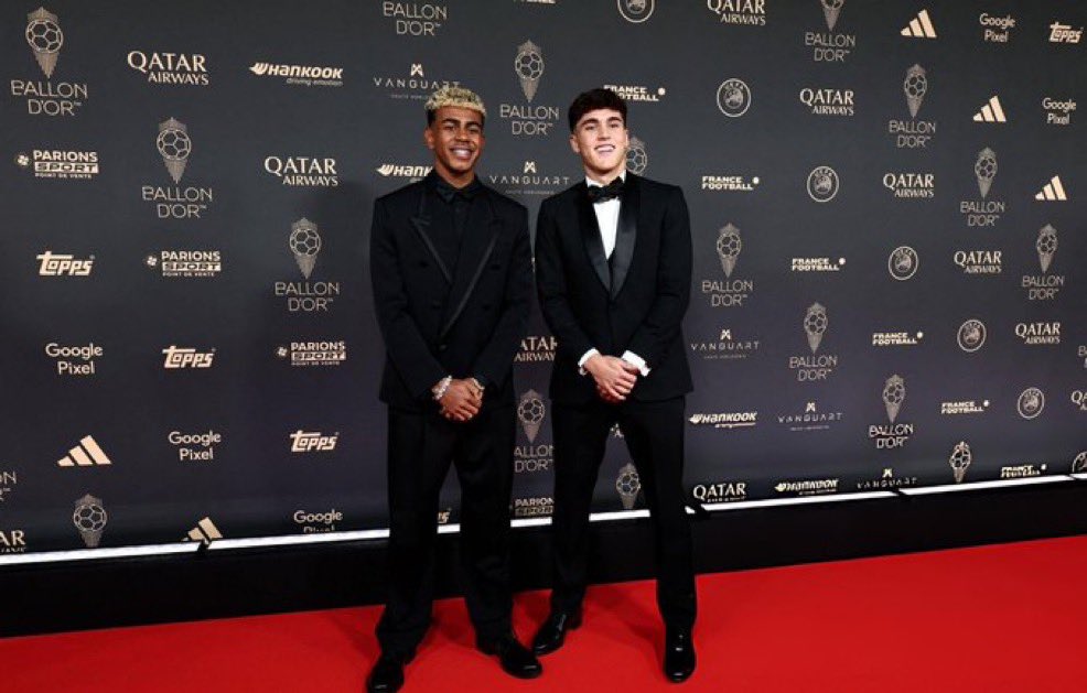 Two men standing on a red carpet, wearing black tuxedos. One has a black shirt and the other a white shirt, both smiling. A backdrop displays logos including Ballon d\'Or, Qatar Airways, Google, and Adidas.