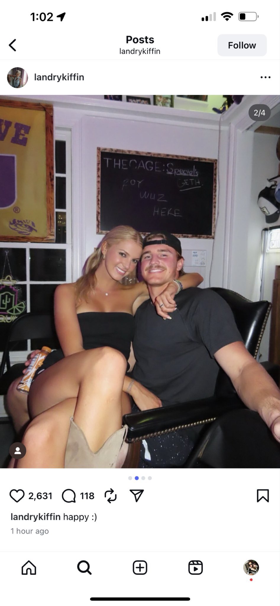 Landry Kiffin and Whit Weeks sitting closely together on a black chair in a room with a chalkboard in the background. Landry Kiffin wears a strapless black top and shorts, while Whit Weeks wears a black shirt and cap. A banner with purple and gold colors is visible on the wall.