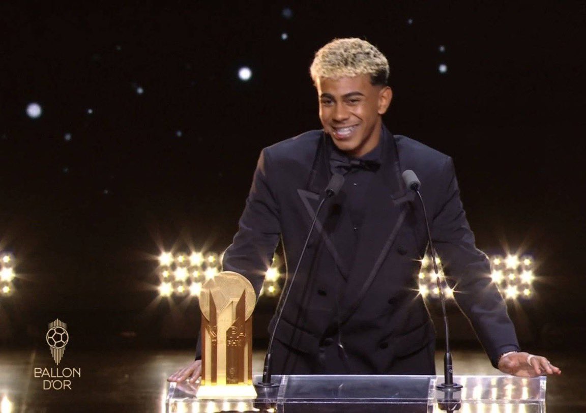 Lamine Yamal standing at a podium, holding a golden trophy. He is wearing a black suit and bow tie, smiling. Bright lights and microphones are visible on the podium.