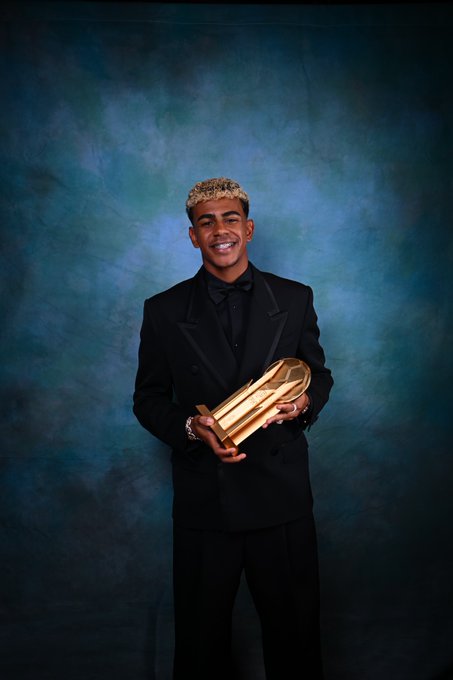 Lamine Yamal stands holding a golden Ballon d\'Or trophy. He wears a black suit, black shirt, and bow tie, with blonde hair. The background is a blue gradient.