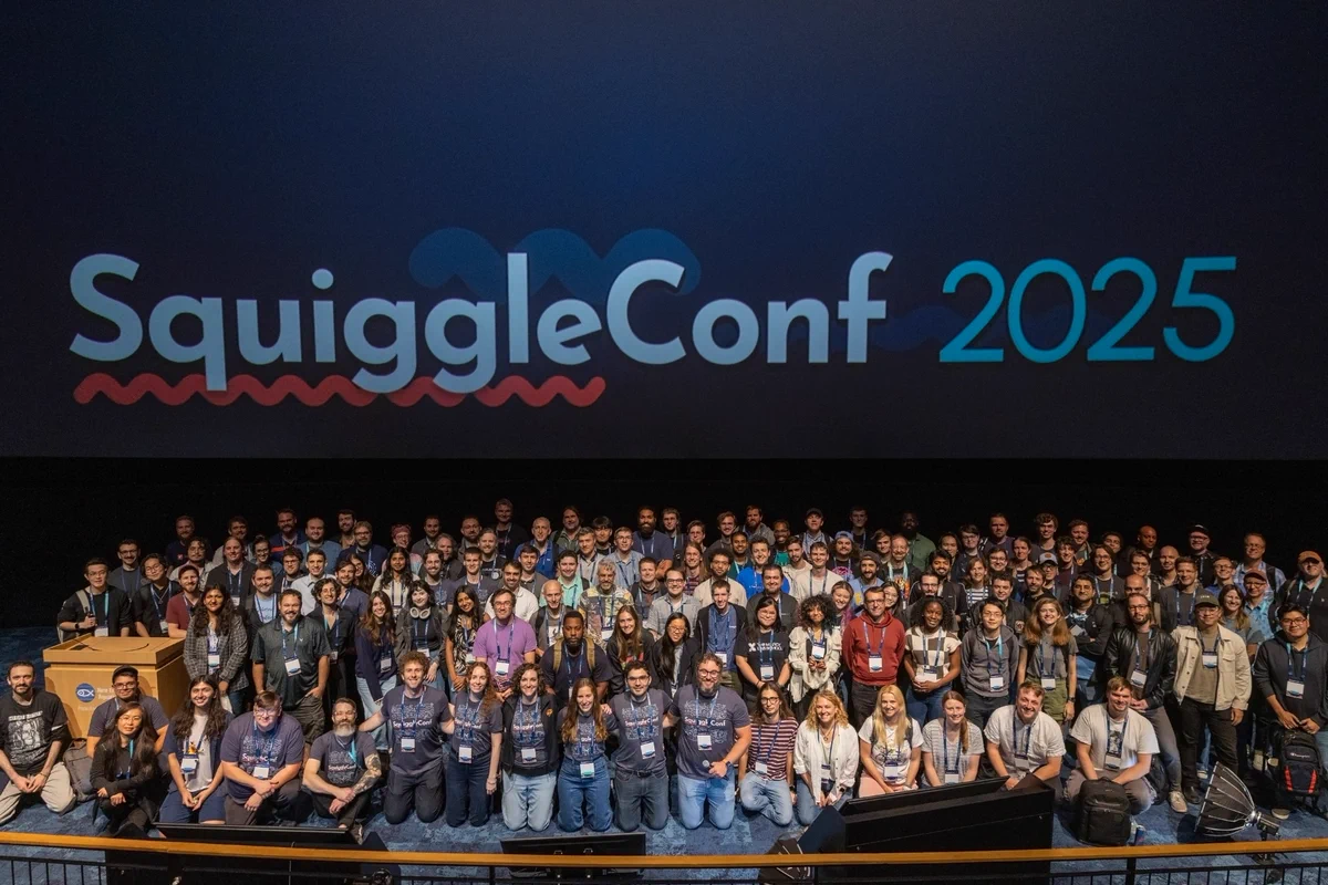 Group photo of smiling attendees, speakers, and organizers on stage with "SquiggleConf 2025" on the aquarium Imax screen behind them.