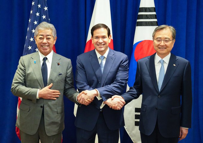 Secretary Rubio stands between Japanese Foreign Minister Takeshi and Republic of Korea Foreign Minister Cho Hyun in New York City on September 22, 2025. The Secretary crosses his arms to shake the hands of each official next to him. U.S., ROK, and Japanese flags are displayed behind the men. (State Department/Freddie Everett)