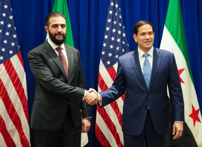 Two men in suits shaking hands in front of American and Syrian flags. One man wears a dark suit with a red tie, the other a blue suit with a blue tie.