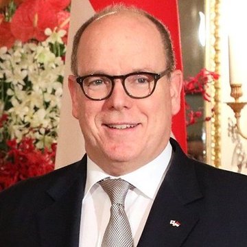 Albert II wearing a dark suit, white shirt, and patterned tie, with a small flag pin on his lapel. Red and white flowers are visible in the background. A flag with red, black, white, and green horizontal stripes is shown.