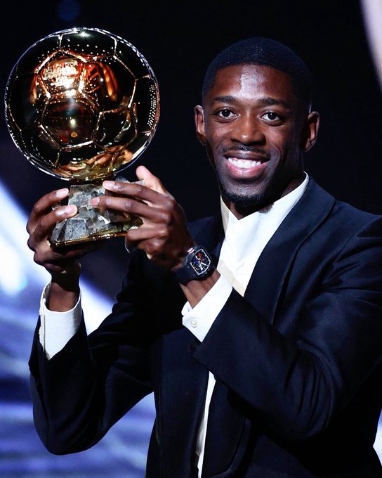 Ousmane Dembélé holding a golden Ballon d’Or trophy with intricate designs. He is wearing a dark suit, white shirt, and a wristwatch. The background is dark with blue lighting.