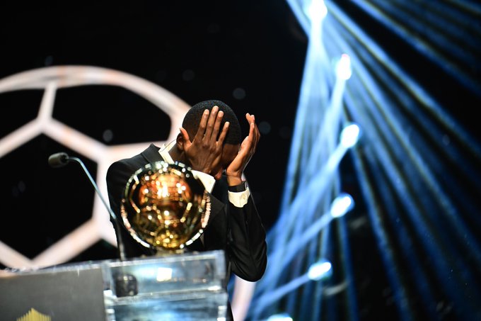 A person in a suit standing at a podium, holding their head with both hands. A golden trophy with a soccer ball design is visible on the podium. Bright lights and a large soccer ball graphic are in the background.