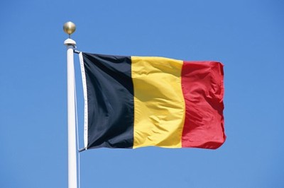 Two flags waving on poles against a blue sky. The first flag has vertical black, yellow, and red stripes, representing Belgium. The second flag has horizontal black, white, and green stripes with a red triangle on the hoist side, representing Palestine.