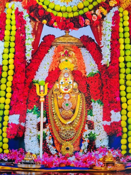 A statue of Bhramaramba Ammavari adorned with gold jewelry and red and white flowers. The statue is decorated with garlands, necklaces, and a crown, standing in a temple setting. Yellow and green garlands frame the statue, and a trident is visible in front.