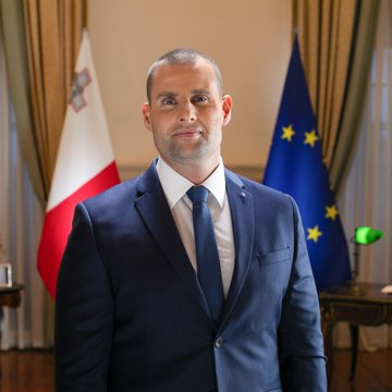 A man in a blue suit and tie stands in a formal room. Behind him are two flags, one with a red and white design and another with a circle of yellow stars on a blue background. A third flag with red, black, white, and green horizontal stripes is also visible.