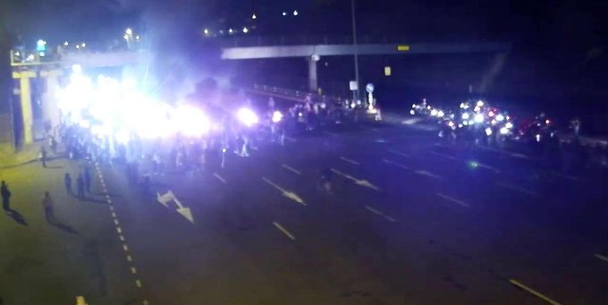 A nighttime scene on a road with multiple burning tires emitting flames and smoke, blocking the street. People stand nearby, some wearing casual clothing like tracksuits. Vehicles, including a truck with visible text, are partially visible. Bright lights from emergency or street sources illuminate the area, with an overpass in the background.