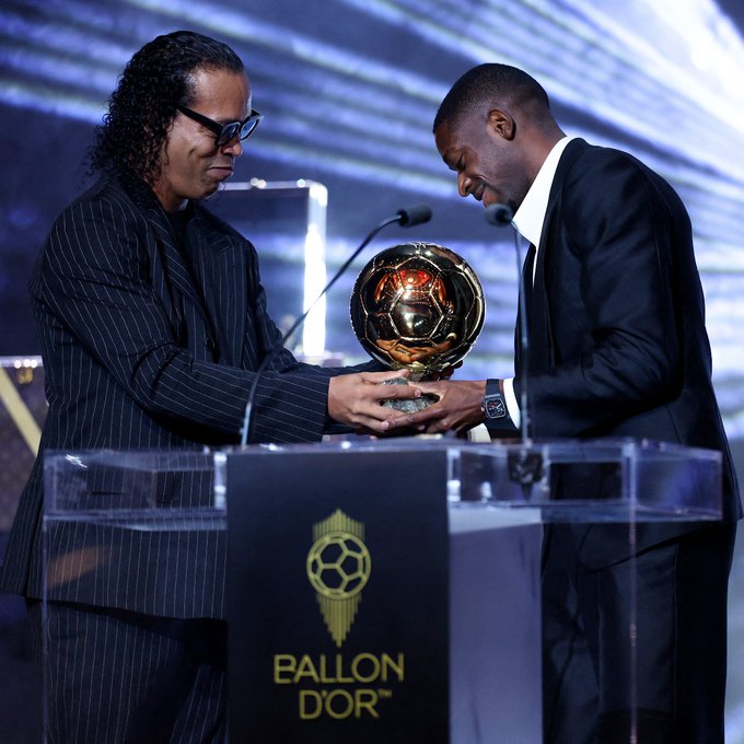 Ronaldinho and Kylian Mbappé standing at a podium. Ronaldinho, with long curly hair and wearing a dark pinstripe suit, holds a golden Ballon d'Or trophy, handing it to Kylian Mbappé, dressed in a black suit. A sign on the podium reads "Ballon d'Or" in gold lettering.