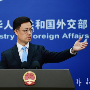 A man in a dark suit and light blue tie stands at a podium with microphones, gesturing with his right hand. The podium bears the emblem of the People\'s Republic of China. Behind him is a blue backdrop with Chinese text reading "中华人民共和国外交部" and English text reading "Ministry of Foreign Affairs." A flag with red, black, white, and green horizontal stripes and a red triangle is visible below.
