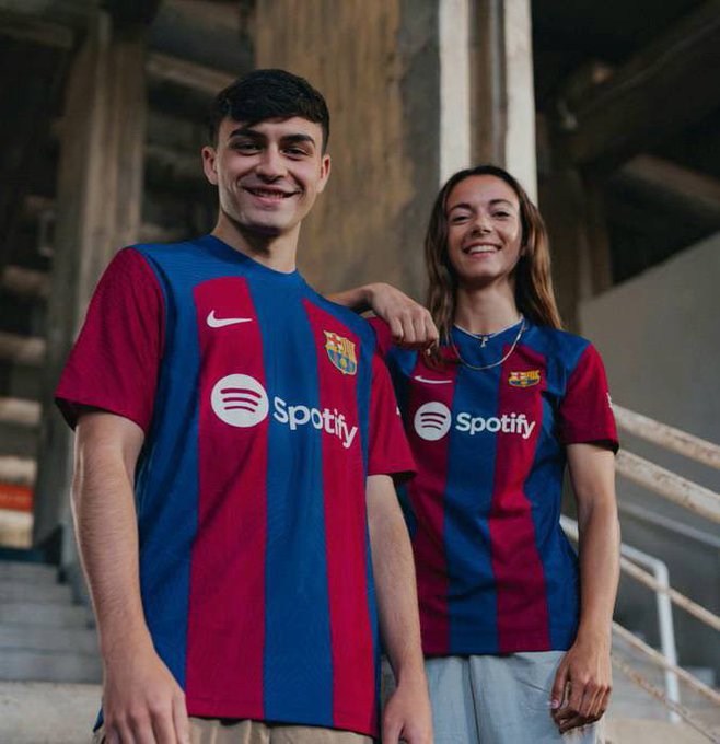 Pedri and Aitana Bonmatí wearing FC Barcelona jerseys with blue and red vertical stripes and the Spotify logo. They stand in front of a concrete structure with stairs and pillars. The jerseys feature the Nike logo and FC Barcelona crest.