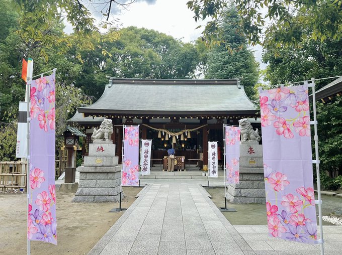 A pathway leading to a traditional Japanese shrine with a tiled roof, flanked by stone statues of guardian lions. Decorative banners with cosmos flower designs in pink and purple stand on either side of the path. Trees and greenery surround the shrine, with a clear sky above.
