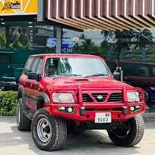 A gray Land Rover and a red Nissan Patrol parked on grass near a building with a "Carnival" sign. Both vehicles have offroad modifications, including large tires and front grilles. License plates are visible on both cars, with text reading "KL 01 DB 6668" on the Land Rover and "KL 01 NF 6663" on the Nissan Patrol.