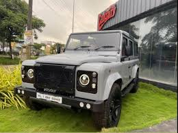 A gray Land Rover and a red Nissan Patrol parked on grass near a building with a "Carnival" sign. Both vehicles have offroad modifications, including large tires and front grilles. License plates are visible on both cars, with text reading "KL 01 DB 6668" on the Land Rover and "KL 01 NF 6663" on the Nissan Patrol.
