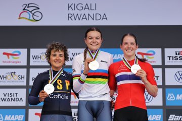 Megan Arens stands in the center wearing a white jersey with rainbow stripes, holding a gold medal and raising her arms. Paula Ostiz Taco is on the left in a blue jersey with "ESP Cofidis" text, holding a silver medal. Oda Aune Gissinger is on the right in a red jersey with Norwegian flag colors, holding a bronze medal. They are on a podium with banners displaying "UCI 2025 World Championships Kigali Rwanda" and sponsor logos like Tissot and Volkswagen.