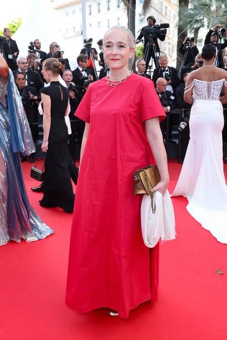 A woman in a long red dress with short sleeves, holding a gold clutch and a white cloth. She stands on a red carpet surrounded by photographers and other people in formal attire, including gowns and suits.