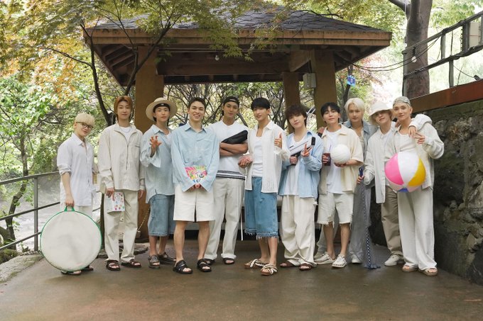 Thirteen individuals, including SEVENTEEN members, standing outdoors on a paved area near a wooden gazebo with a sloped roof. They wear light-colored casual outfits, some in white shirts and shorts, others in light blue shirts and pants, with sandals or sneakers. Several hold recreational items like a beach ball, a large circular object, and papers, suggesting a summer outing.