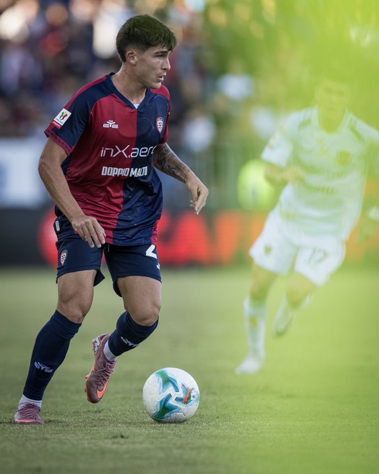 A soccer player in a red and blue Cagliari Calcio jersey with the number 24, running on a grassy field with a soccer ball at his feet. He wears pink cleats and dark socks. Another player in a white jersey is visible in the background, slightly blurred.