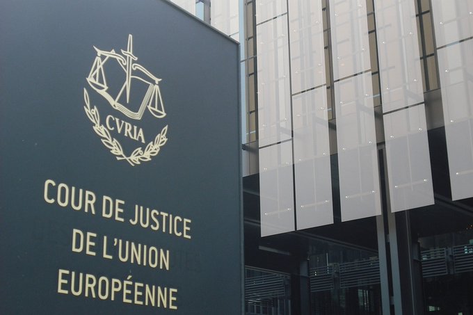 A sign with a gold emblem featuring scales, a cross, and laurel branches, labeled "CURIA" above the text "COUR DE JUSTICE DE L'UNION EUROPÉENNE" in gold letters on a dark background. The sign is mounted on a building with a modern facade featuring vertical slats and glass windows.