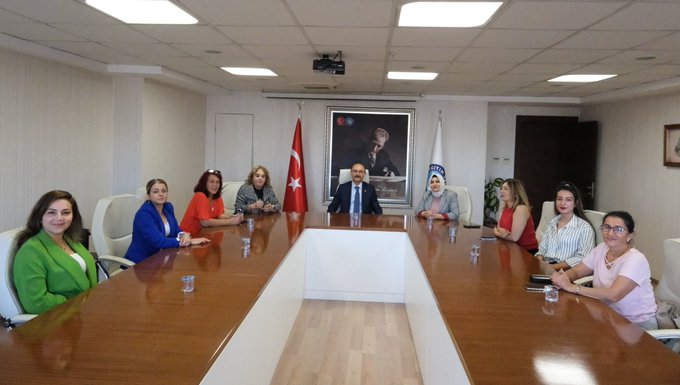 A group of people seated around a long wooden table in a meeting room. Turkish flags and a banner with a blue emblem and text "Eğitim 1992" are visible behind them. A large framed portrait of a man is on the wall. Some individuals wear formal attire, including suits and blazers in various colors like blue, green, and red.