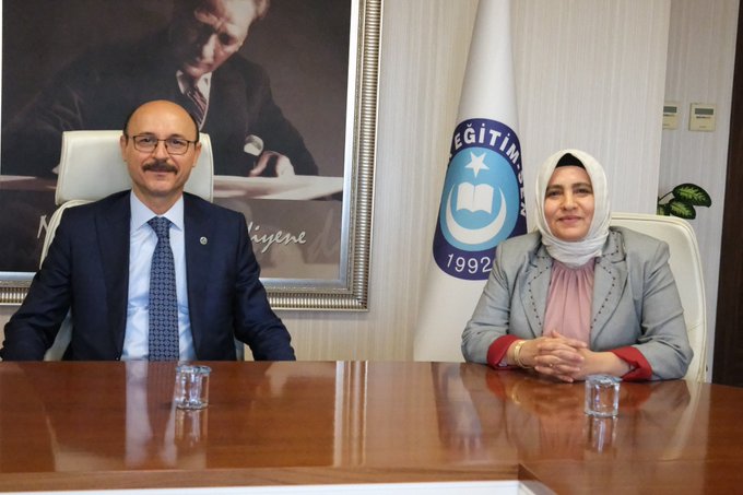 A group of people seated around a long wooden table in a meeting room. Turkish flags and a banner with a blue emblem and text "Eğitim 1992" are visible behind them. A large framed portrait of a man is on the wall. Some individuals wear formal attire, including suits and blazers in various colors like blue, green, and red.