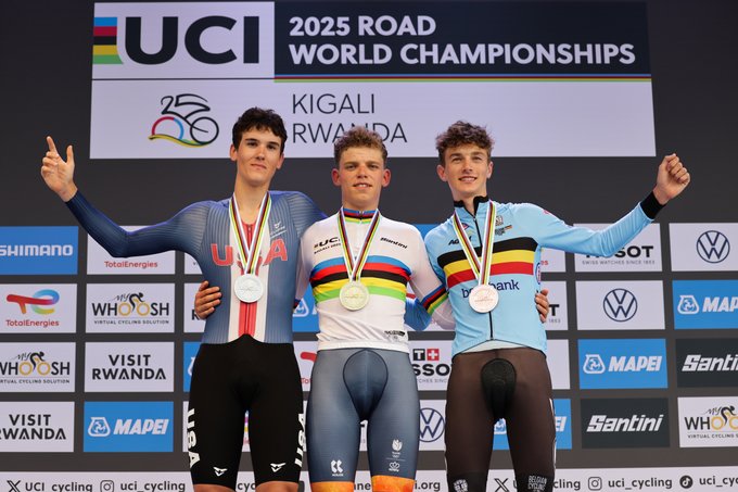 Michiel Mouris, Ashlin Barry, and Seff van Kerckhove standing on a podium wearing cycling uniforms. Michiel Mouris in the center wears a rainbow-striped jersey and a gold medal. Ashlin Barry on the left wears a blue and red uniform with a silver medal. Seff van Kerckhove on the right wears a blue and yellow uniform with a bronze medal. A backdrop displays "2025 UCI Road Championships Kigali Rwanda" and logos including Shimano, VW, and Mapei.