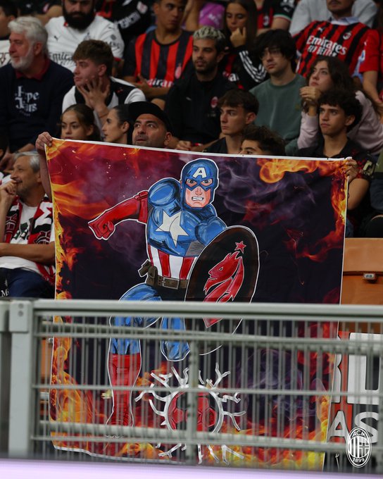 A crowd of people at a soccer match, many wearing red and black AC Milan jerseys. One person holds a large flag featuring Captain America in a dynamic pose, with a red and black design and flames in the background. A metal barrier is visible in the foreground.