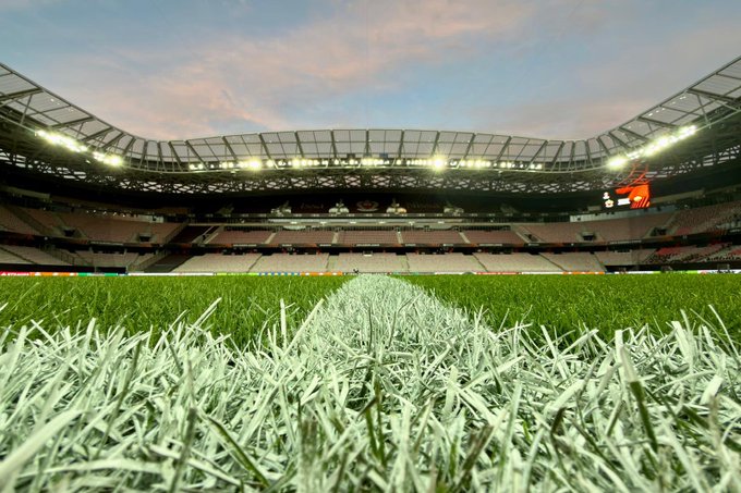 A large soccer stadium with a well-lit field at dusk. The grass is green and neatly striped, extending toward the stands. The stands are mostly empty, with rows of seats visible under a curved, translucent roof. Bright stadium lights illuminate the field and upper structure.