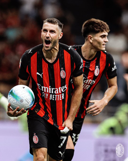 Two men wearing black and red striped jerseys with "Emirates Fly Better" text, running on a field. One man holds a soccer ball, wearing a jersey with the number 7. The jerseys display the AC Milan logo.