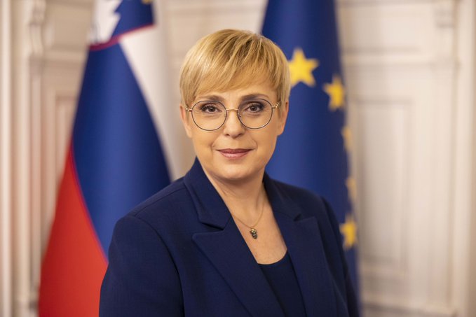 A woman with short blonde hair and glasses, wearing a blue blazer and necklace, standing in front of the Slovenian flag and the European Union flag. A flag with red, black, white, and green horizontal stripes, featuring a red triangle on the left side.