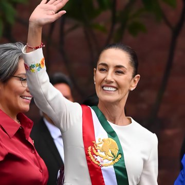 Claudia Sheinbaum waving with her right hand raised, wearing a white outfit with a green, white, and red sash featuring the Mexican flag and eagle emblem. A bracelet with colorful floral designs is on her wrist. Another person in a red shirt is partially visible, smiling beside her, with a brick wall and greenery in the background. A second image shows the Palestinian flag with red, black, white, and green horizontal stripes.