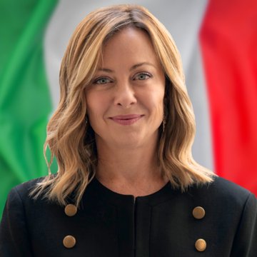 A woman with shoulder-length blonde hair wearing a black jacket with gold buttons stands in front of the Italian flag, featuring green, white, and red vertical stripes. A naval ship with a similar Italian flag flies on its mast, sailing on the ocean with another ship visible in the background under a cloudy sky.