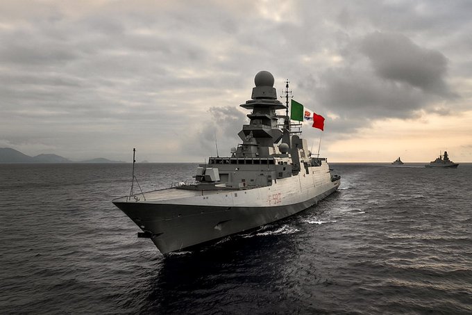 A woman with shoulder-length blonde hair wearing a black jacket with gold buttons stands in front of the Italian flag, featuring green, white, and red vertical stripes. A naval ship with a similar Italian flag flies on its mast, sailing on the ocean with another ship visible in the background under a cloudy sky.