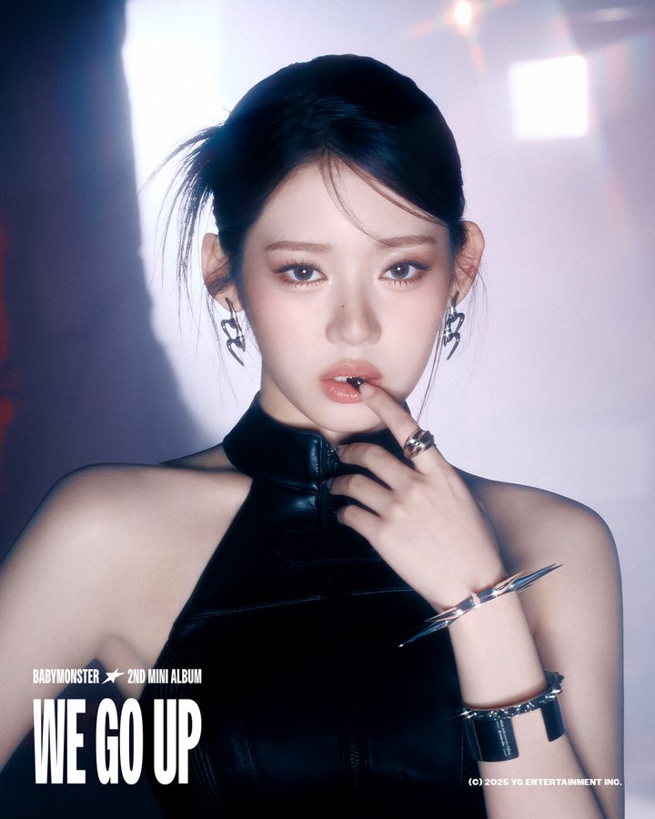 Rora from BABYMONSTER, wearing a black sleeveless top with a high neckline and metallic accessories including rings, bracelets, and earrings shaped like lightning bolts. Her hair is styled in a high ponytail. The background features soft lighting with bokeh effects and a blurred urban setting.