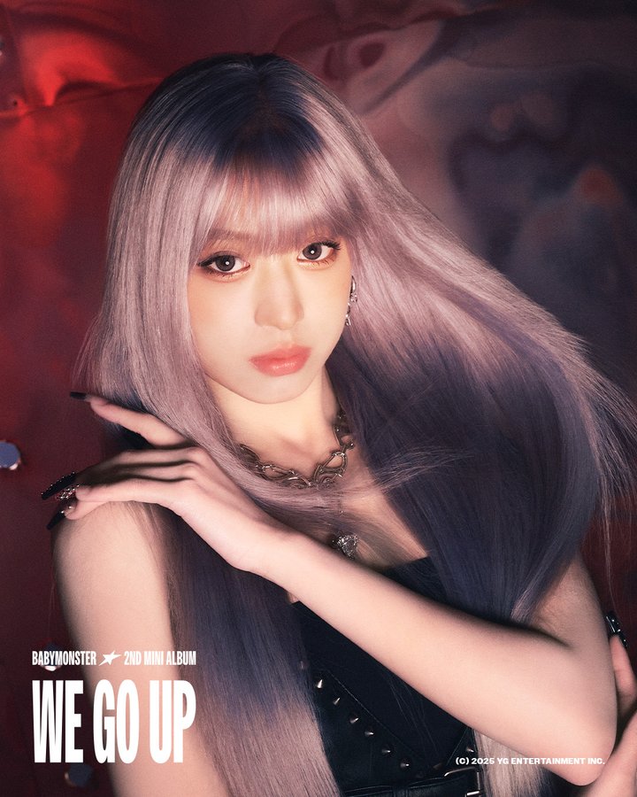 Ahyeon with long, straight, light-colored hair, wearing a black outfit and a necklace. She poses with one hand near her face, against a red and abstract background.