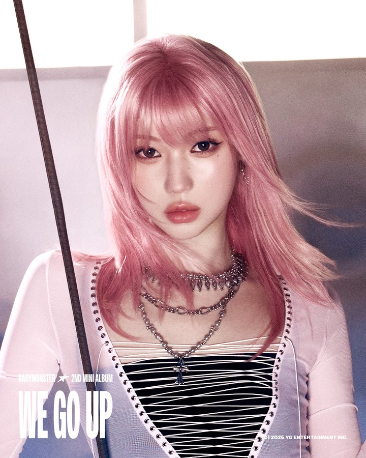 Asa with pink hair wearing a light pink cardigan over a black and white striped top. She has multiple layered necklaces with metallic chains and pendants. A riding crop is positioned diagonally across her chest.