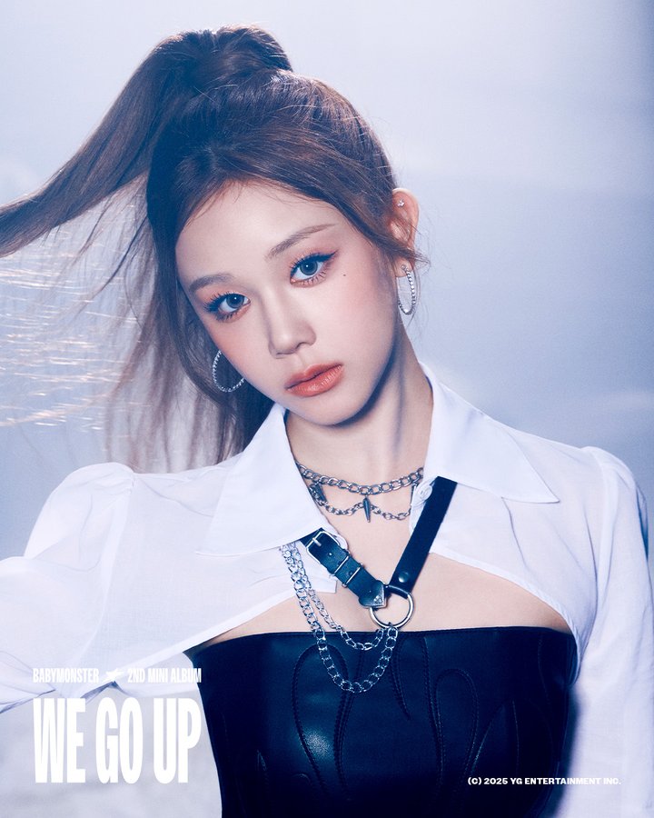 Pharita from BABYMONSTER, wearing a white shirt over a black top with chain accessories and hoop earrings. Her hair is styled in a high ponytail. Text overlay reads