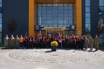 A group of students holding Turkish flags stands in front of a building with a sign reading "Kara Kuvvetleri Hava Savunma Komutanlığı Eğtim ve Simülasyon Merkezi." Military personnel in uniform are present. Another image shows students with Turkish flags observing military equipment, including a missile launcher and armored vehicles, with a person in a safety vest giving a presentation. A third image depicts students holding Turkish flags watching a military tank in an open field.