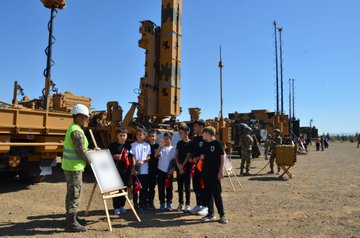 A group of students holding Turkish flags stands in front of a building with a sign reading "Kara Kuvvetleri Hava Savunma Komutanlığı Eğtim ve Simülasyon Merkezi." Military personnel in uniform are present. Another image shows students with Turkish flags observing military equipment, including a missile launcher and armored vehicles, with a person in a safety vest giving a presentation. A third image depicts students holding Turkish flags watching a military tank in an open field.