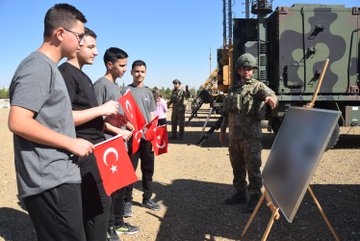 A group of students holding Turkish flags stands in front of a building with a sign reading "Kara Kuvvetleri Hava Savunma Komutanlığı Eğtim ve Simülasyon Merkezi." Military personnel in uniform are present. Another image shows students with Turkish flags observing military equipment, including a missile launcher and armored vehicles, with a person in a safety vest giving a presentation. A third image depicts students holding Turkish flags watching a military tank in an open field.