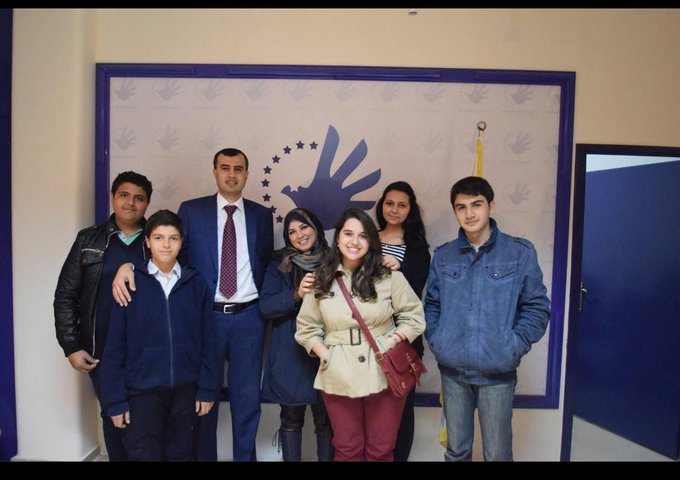A group of seven people, including Ghada Rabah, standing together indoors. They are dressed in casual and formal clothing, with Ghada Rabah in a beige coat and red pants. A blue and white logo with handprints and stars is visible on the wall behind them.
