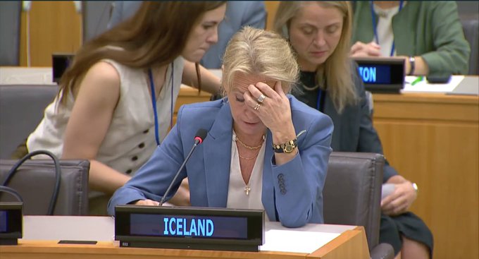 A woman with blonde hair wearing a blue blazer and white shirt sits at a desk, hand on forehead, appearing emotional. A nameplate reading "ICELAND" is in front of her. Other individuals are seated at desks in the background.