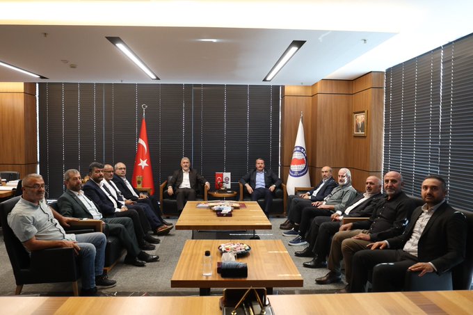 Two groups of men in a meeting room. One group sits at a table with Turkish and white flags with blue circular emblems behind them. The other group stands holding a poster with text and imagery of a raised fist breaking chains, featuring the Palestinian and Turkish flags. The poster includes text in Turkish.