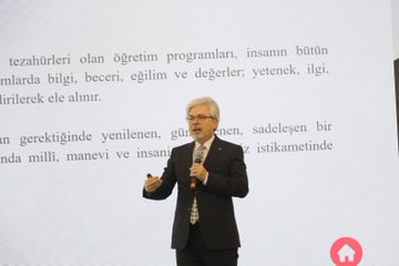 A man in a suit speaking at a podium, holding a microphone. Behind him, a red backdrop displays the logo and text of the Turkish Ministry of National Education and "Türkiye Yüzyılı Maarif Modeli." Rows of men in suits sit in chairs, facing the speaker. Bottles of water and glasses are on tables in front of the seated men. An audience is visible in the background, seated in rows of red chairs.