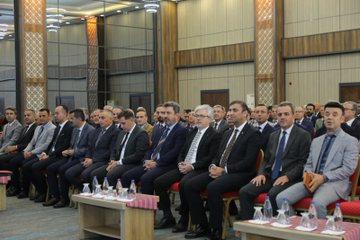 A man in a suit speaking at a podium, holding a microphone. Behind him, a red backdrop displays the logo and text of the Turkish Ministry of National Education and "Türkiye Yüzyılı Maarif Modeli." Rows of men in suits sit in chairs, facing the speaker. Bottles of water and glasses are on tables in front of the seated men. An audience is visible in the background, seated in rows of red chairs.