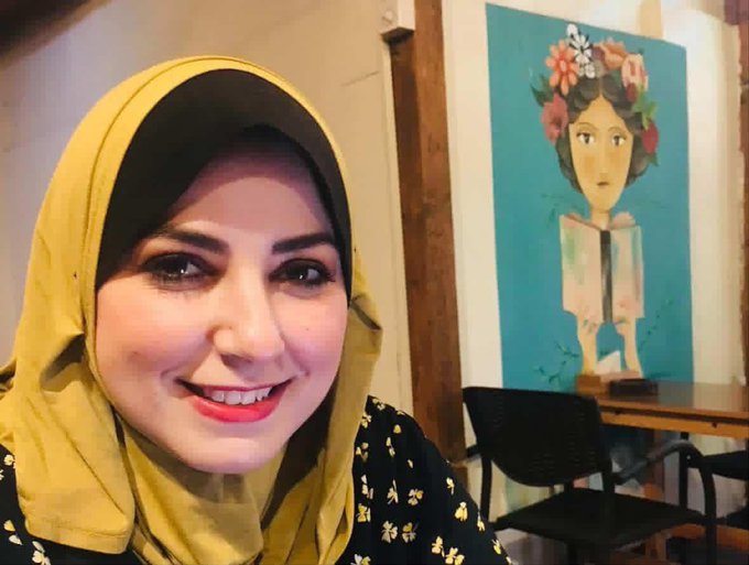 A woman wearing a yellow hijab and black floral dress sits indoors, smiling, with a colorful mural of a woman with flowers in her hair on the wall behind her. A damaged multi-story building with rubble and debris in front, partially collapsed, visible on a phone screen held by a hand.