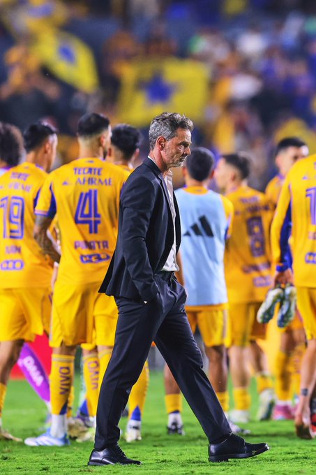 Diego Cocca walking on a soccer field, wearing a black suit and white shirt. Behind him, Tigres players in yellow and purple jerseys with numbers like 19 and 44 visible, standing on the grass.