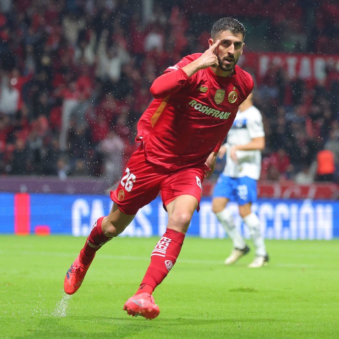 Alexis Vega in a red Toluca uniform, number 25, running on a soccer field, pointing with one finger. Paulinho, also in a red Toluca uniform, is visible in the background. The field has green grass, and a crowd in red fills the stands. Text on the field reads "CÁPSULAS."